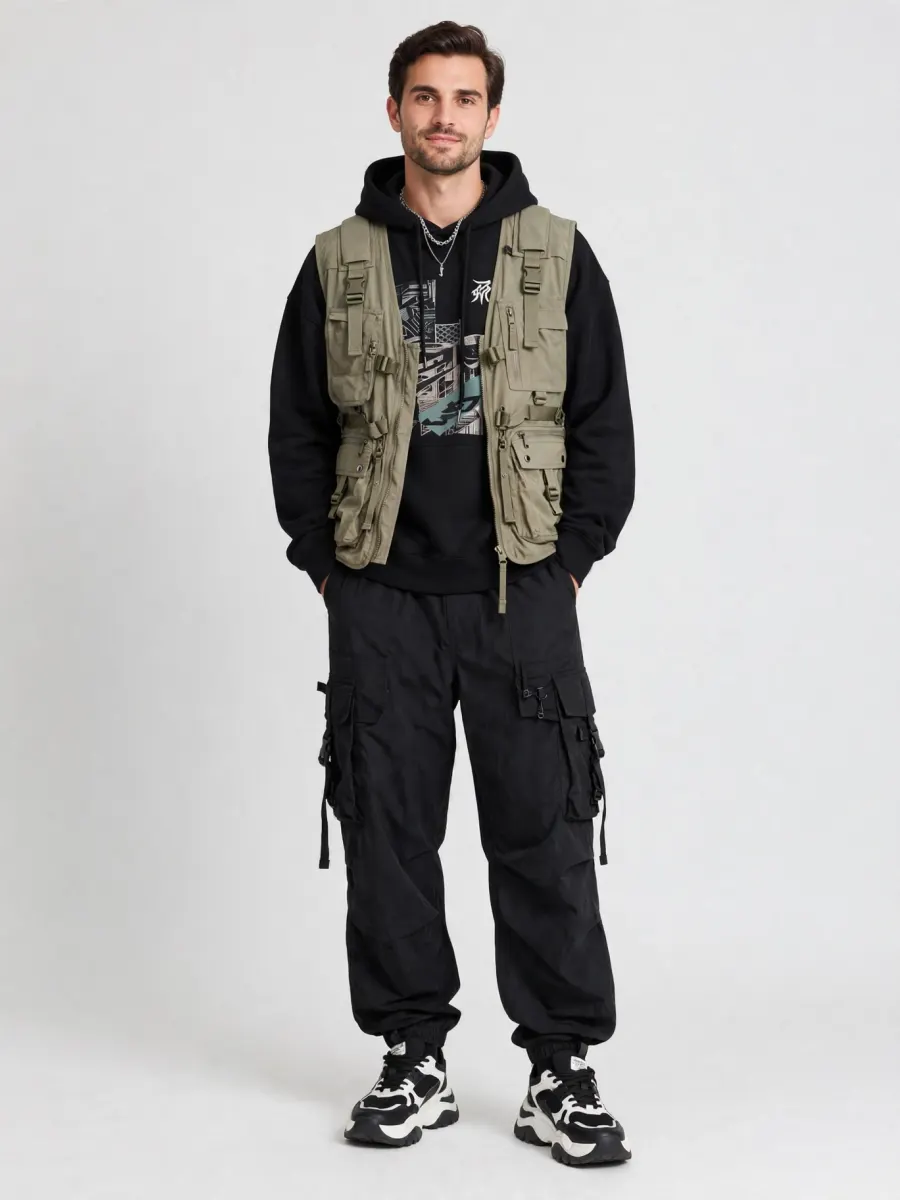 The same man in a black hoodie under a khaki utility vest, black cargo pants and chunky sneakers